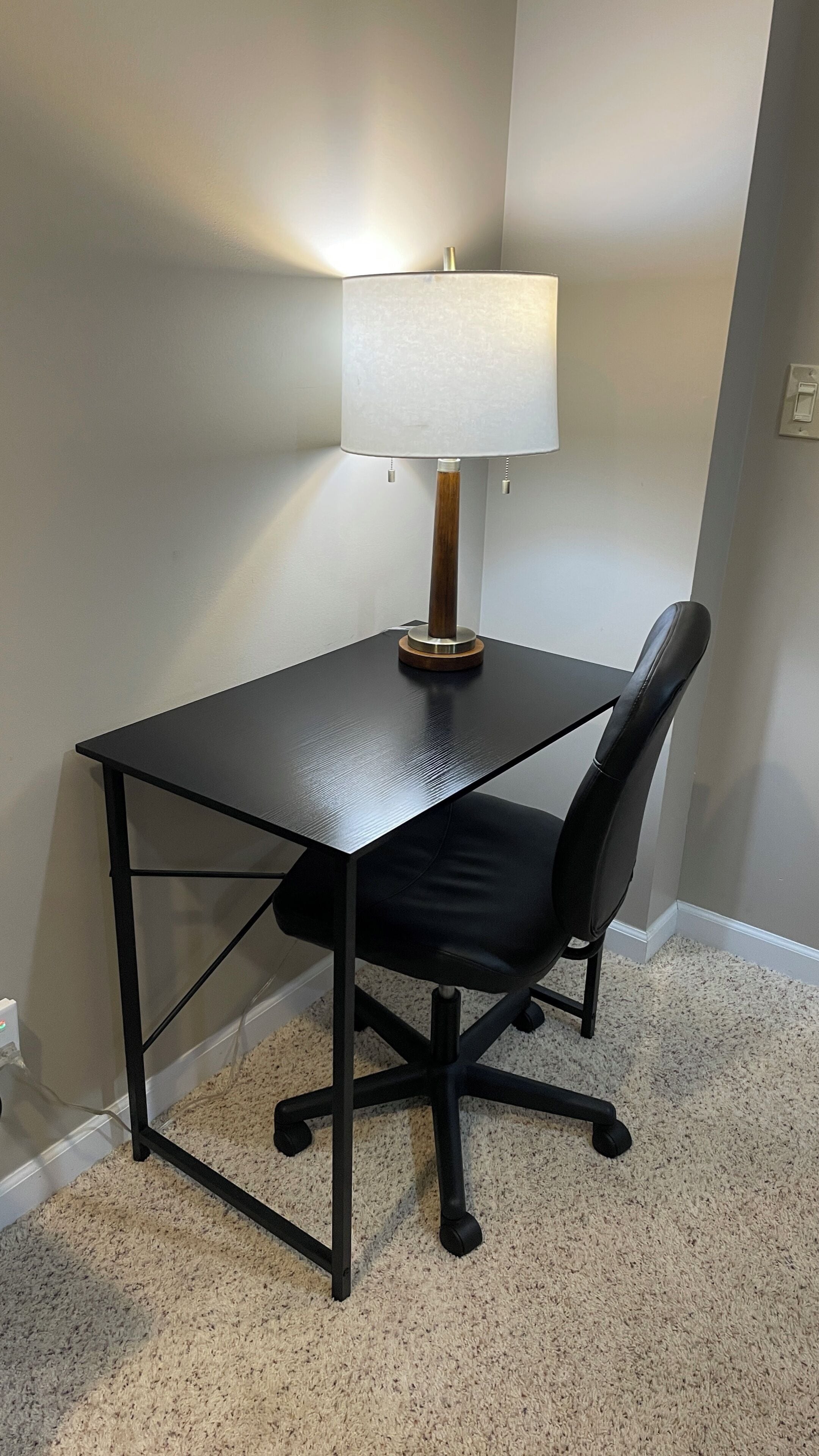 A small, dark-colored desk with a white lamp and a black office chair in a room with a carpeted floor.