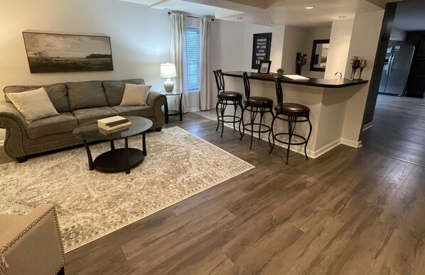Un salon comprenant un canapé, une table basse et un tapis à motifs. On y trouve également un coin repas avec un bar et des tabourets.