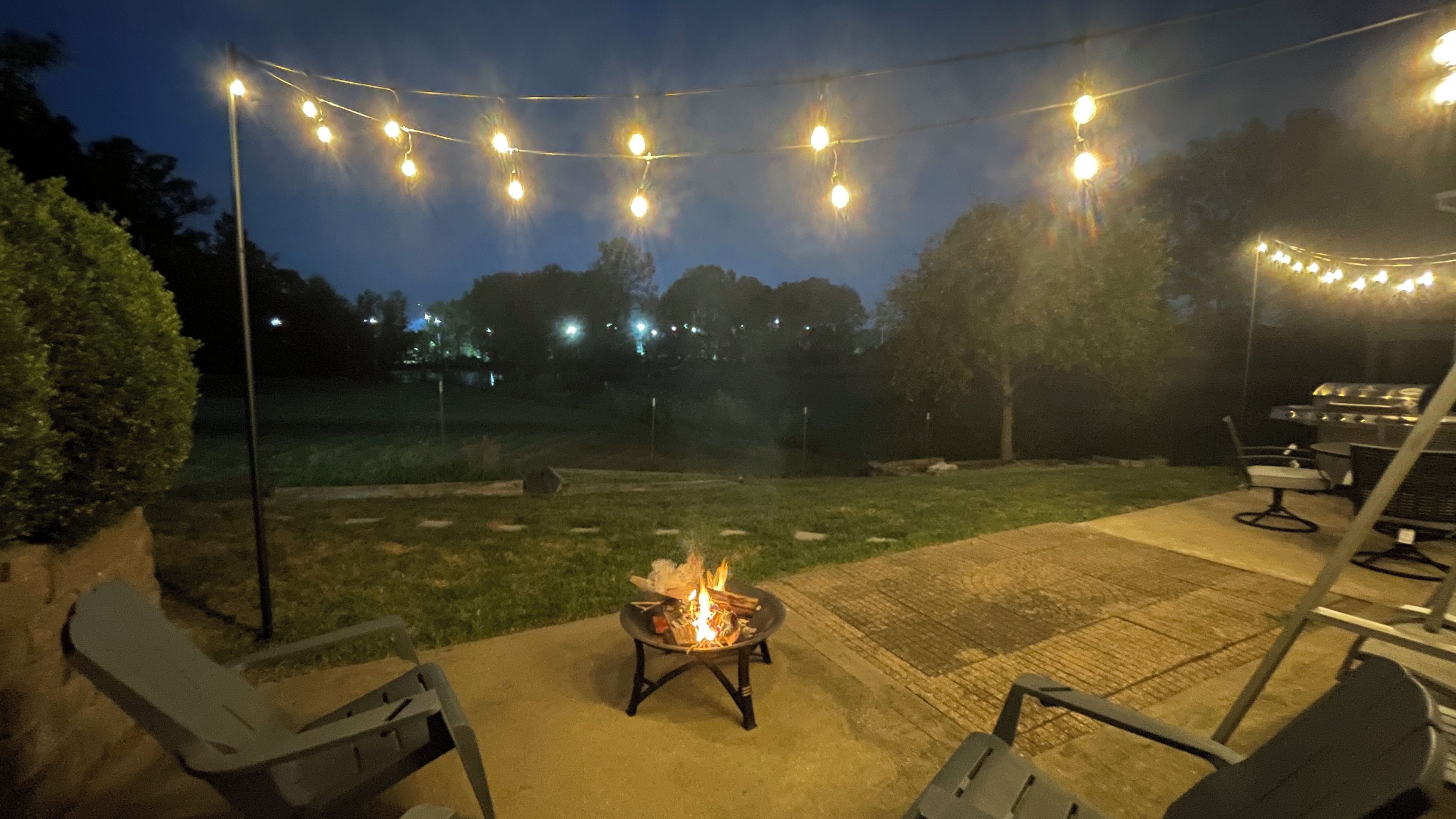 Een terras buiten met een vuurkuil, lichtjes en tuinmeubilair.