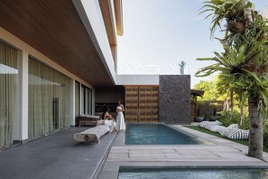 Un área de piscina moderna al aire libre con un deck de madera, una pared de piedra y un espacio de descanso con cojines a rayas.