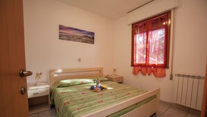 A bedroom with a bed, a nightstand, a bedside table, a window with curtains, a radiator, and a painting on the wall. - Charming 2-room Apartment in a Peaceful Residential Area (Rosolina)