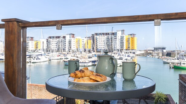 Un balcon avec une table préparée pour le petit-déjeuner, donnant sur une marina où l’on peut admirer des bateaux et des bâtiments.