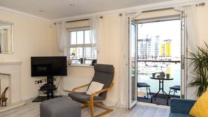 Un salon équipé d’un téléviseur, d’un canapé gris, d’une chaise en bois et d’un balcon offrant une vue sur la ville et l’eau.