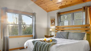 A bedroom with a bed, two windows, a nightstand, a lamp, and a painting on the wall. - Mountaintop Cabin with Panoramic Views, Fire Pit (Waynesville)