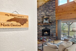 Une enseigne en bois représentant une carte de la Caroline du Nord et portant l’inscription : « Mon cœur est en Caroline du Nord ». Un salon chaleureux avec une cheminée en pierre, un canapé et une table basse.