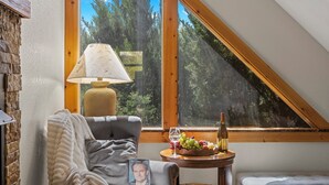 Une pièce chaleureuse avec un plafond en pente, une fenêtre donnant sur les arbres, une lampe, une table avec une bouteille et des fruits, et une chaise avec une couverture.