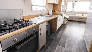 Cocina compacta con estufa a gas, horno y lavaplatos. Hay una encimera de madera y una ventana con persianas. Se ve un sofá y una pequeña mesa en la zona de estar contigua.