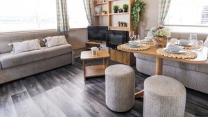 Una sala moderna con un sofá, otomanas, un televisor, una mesa de madera dispuesta para comer y una estantería con plantas.