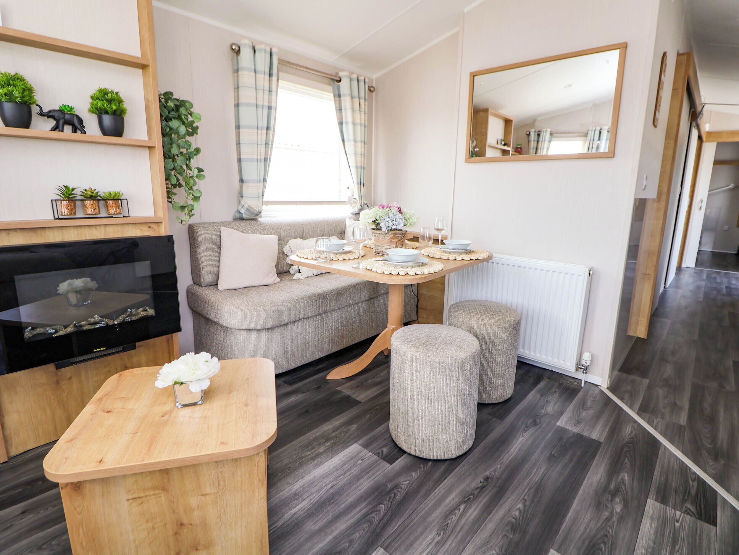 A cozy room with a sofa, a wooden table, and a TV. There are plants on shelves and a window with curtains.