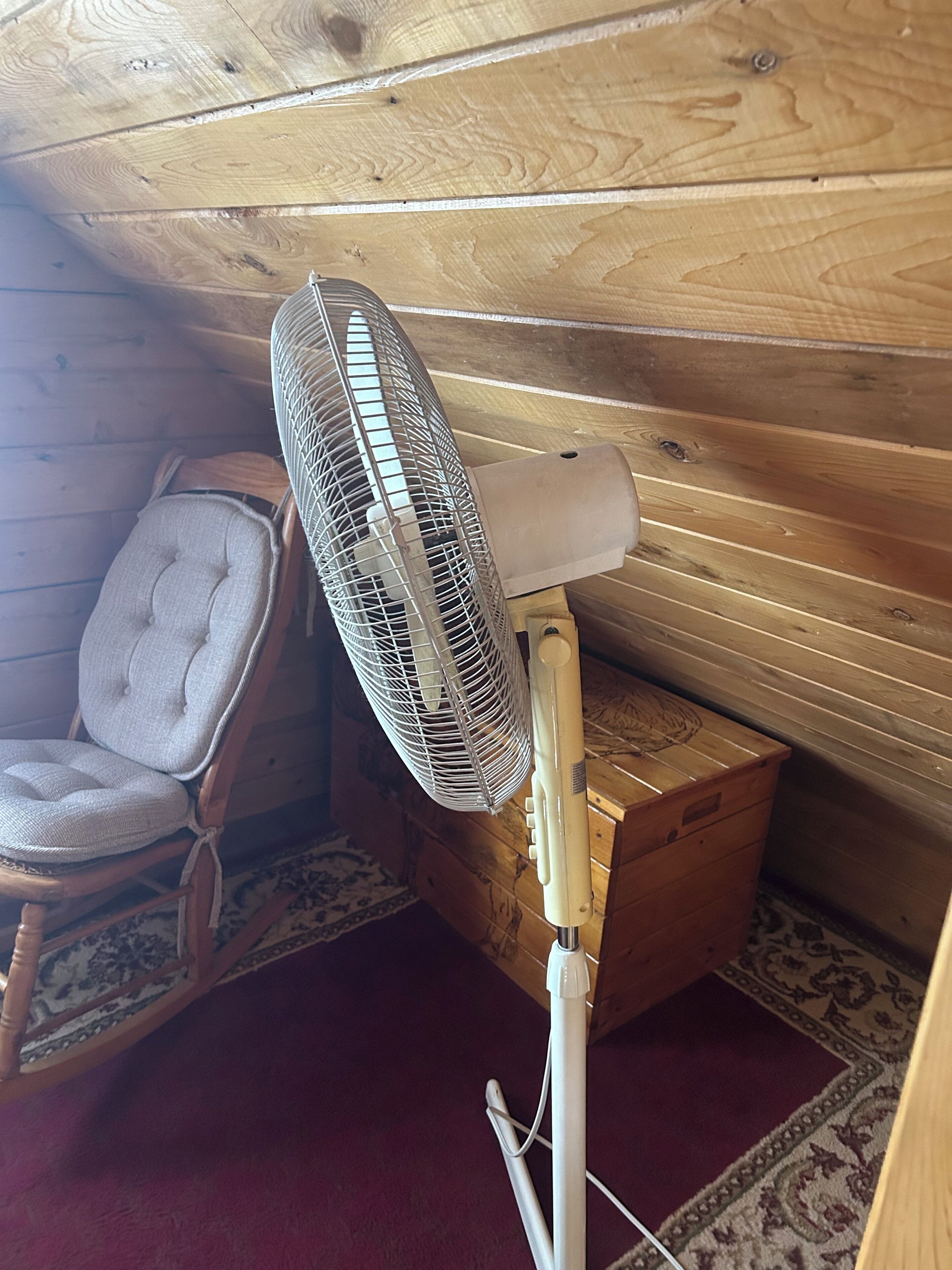Una habitación de madera con un ventilador de pie, un sillón acolchado y una alfombra estampada.