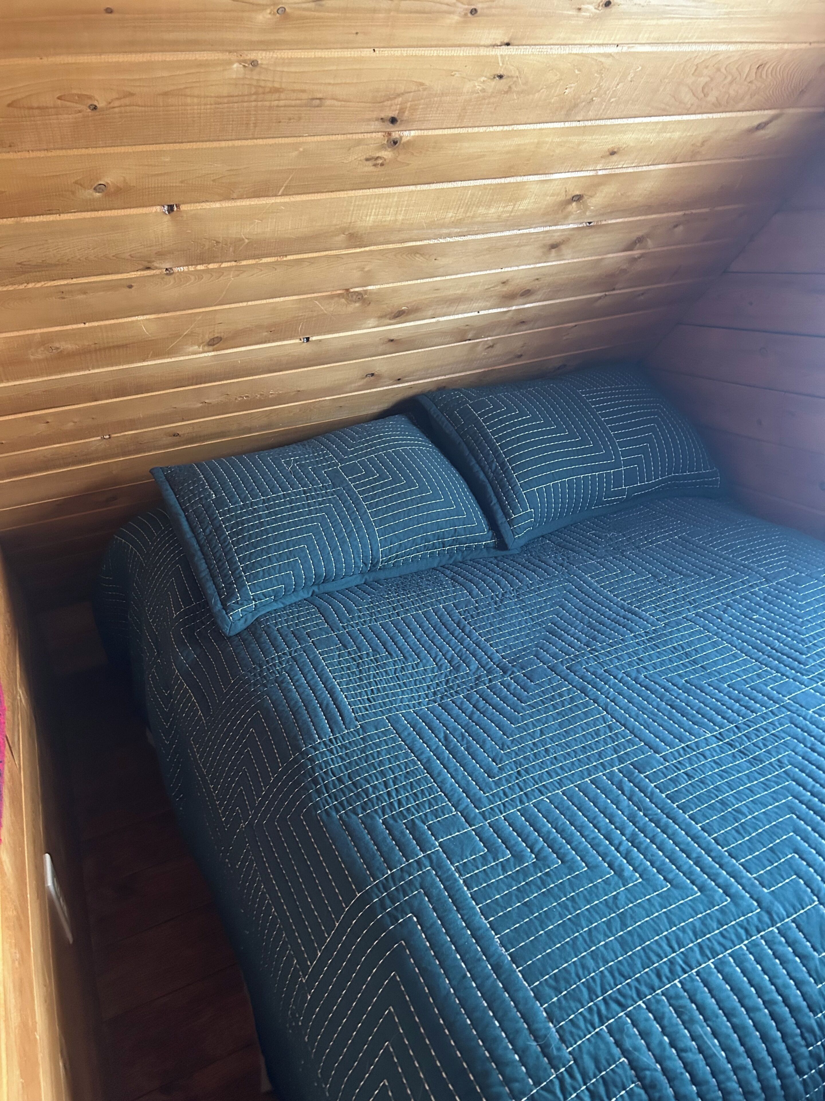 A wooden cabin interior with a bed featuring a blue quilted bedspread and pillows.