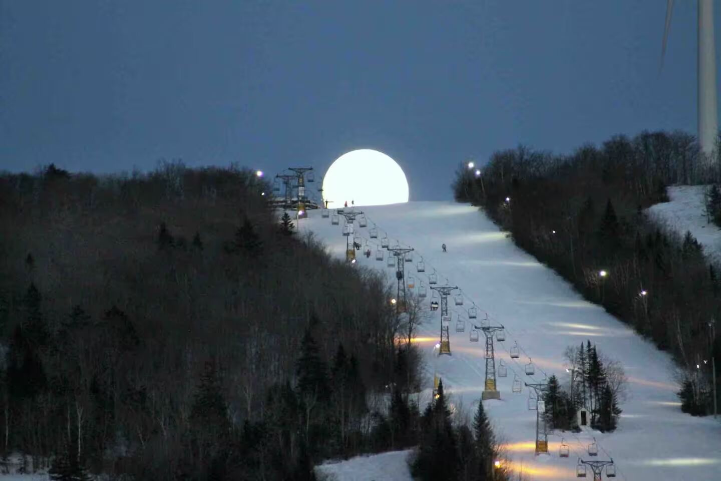 A snow-covered ski slope with a large moon in the sky, surrounded by trees and ski lifts.