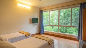 Une chambre d’hôtel avec deux lits, un ventilateur de plafond, une télévision et une grande fenêtre donnant sur la forêt.