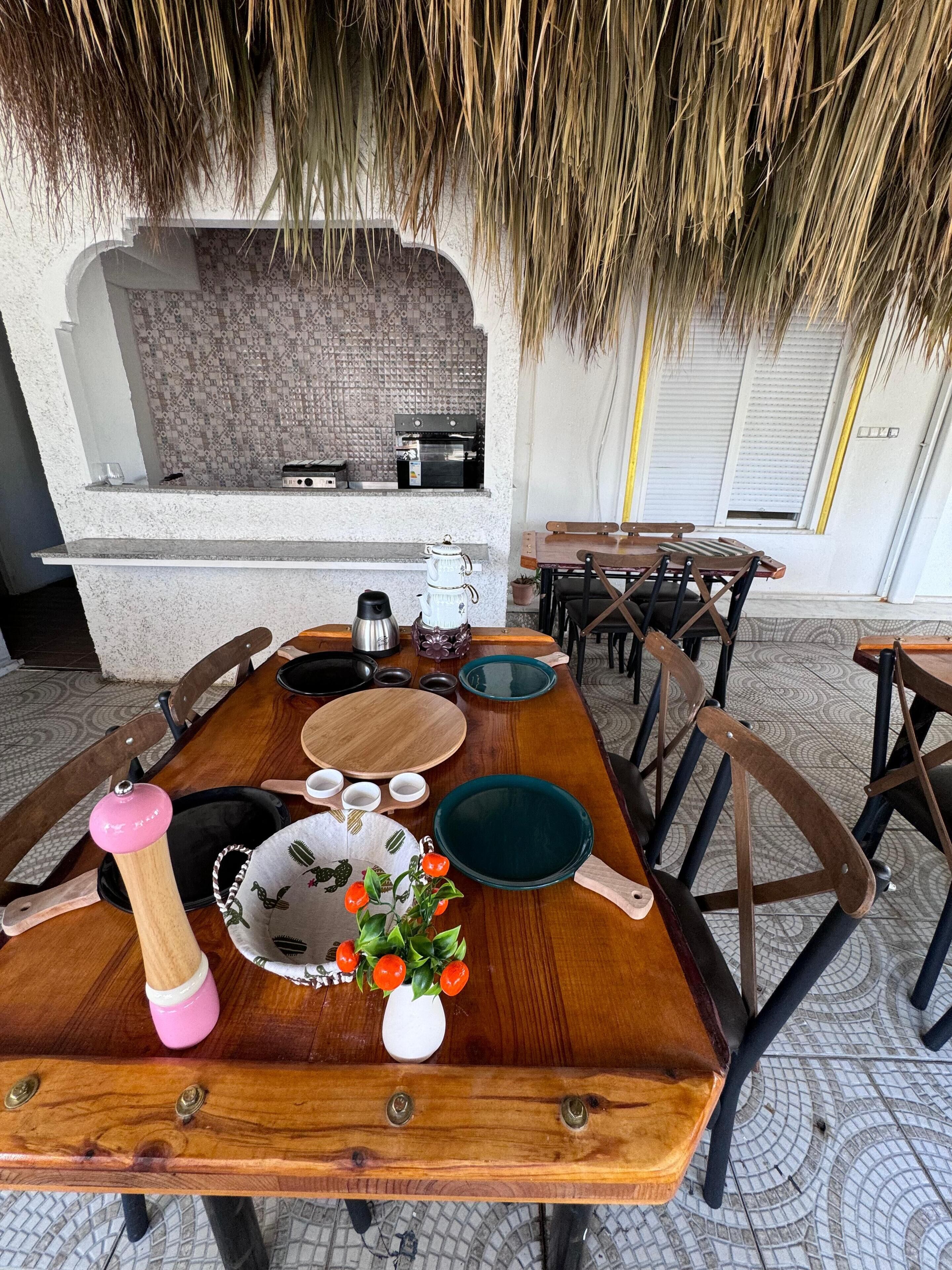 Un comedor con una mesa de madera dispuesta para cuatro personas, una máquina de café y un jarrón decorativo con flores rojas.