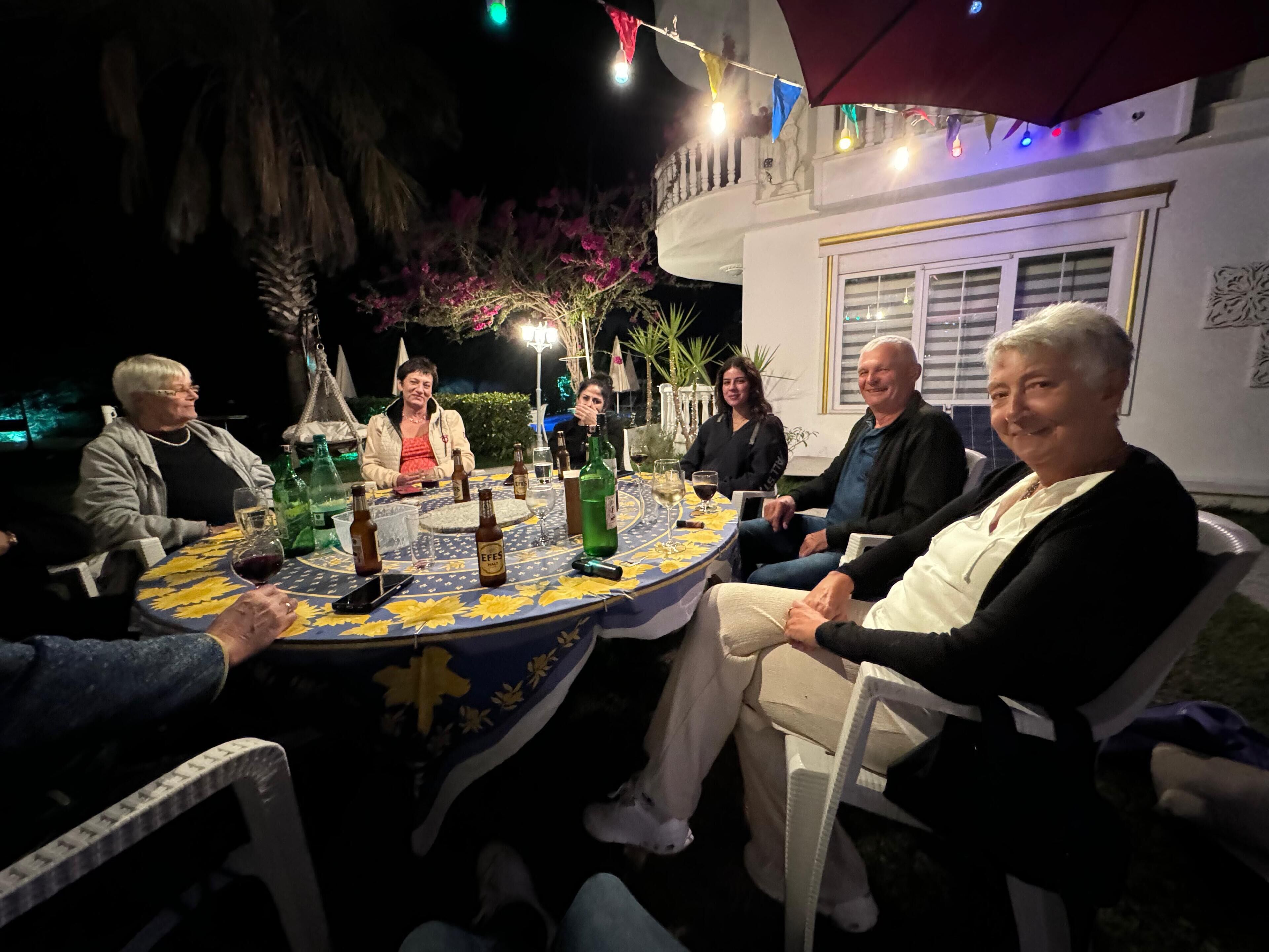 Un grupo de personas sentadas alrededor de una mesa redonda con mantel floral, bajo un espacio al aire libre techado con luces colgantes.