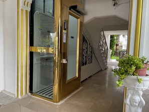L’entrée d’un hôtel, avec une porte dorée, un ascenseur en verre et un escalier orné de balustrades travaillées.
