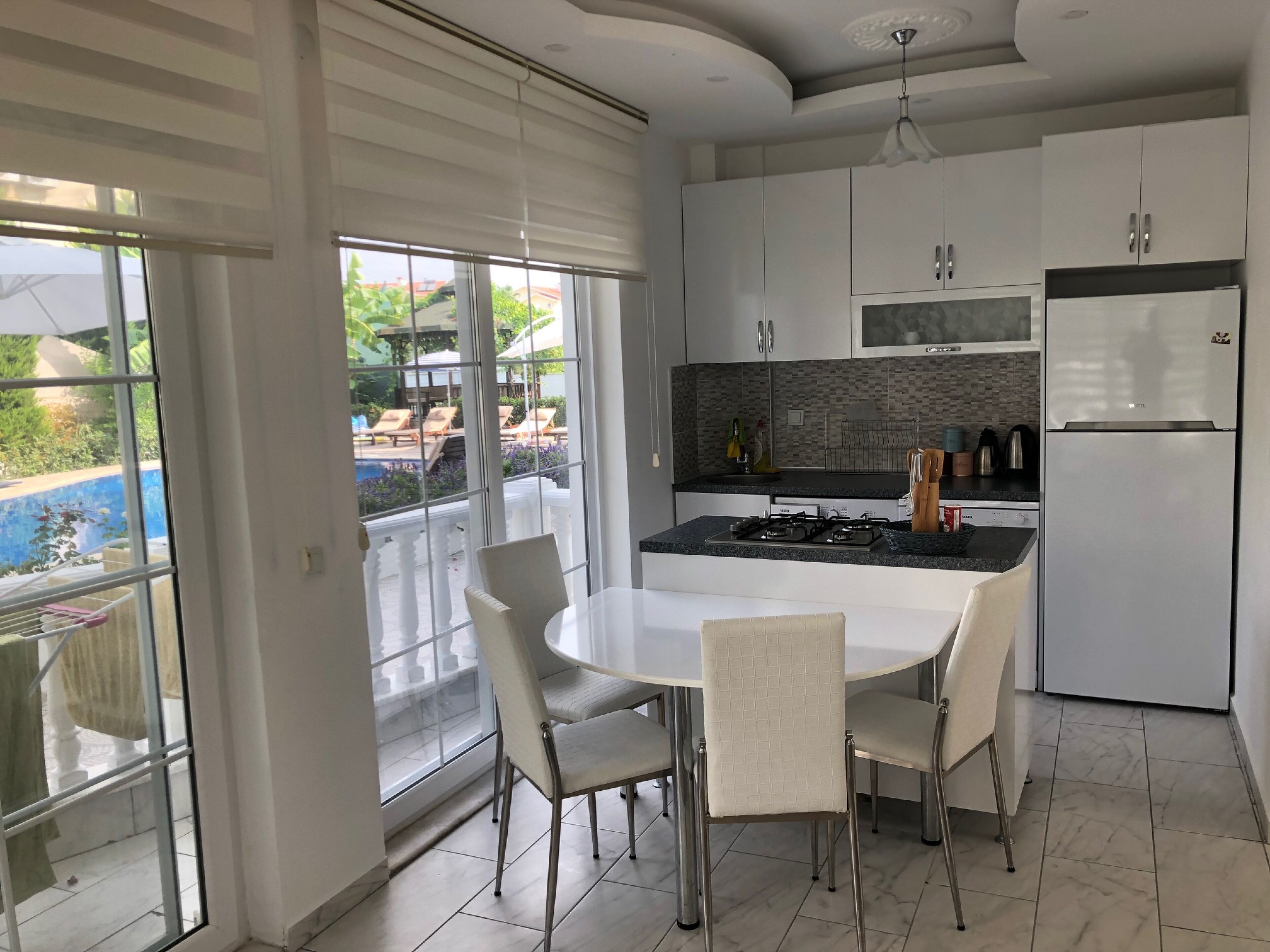 Une cuisine moderne dotée d’un îlot central, de meubles blancs et offrant une vue sur une piscine grâce à de grandes fenêtres.