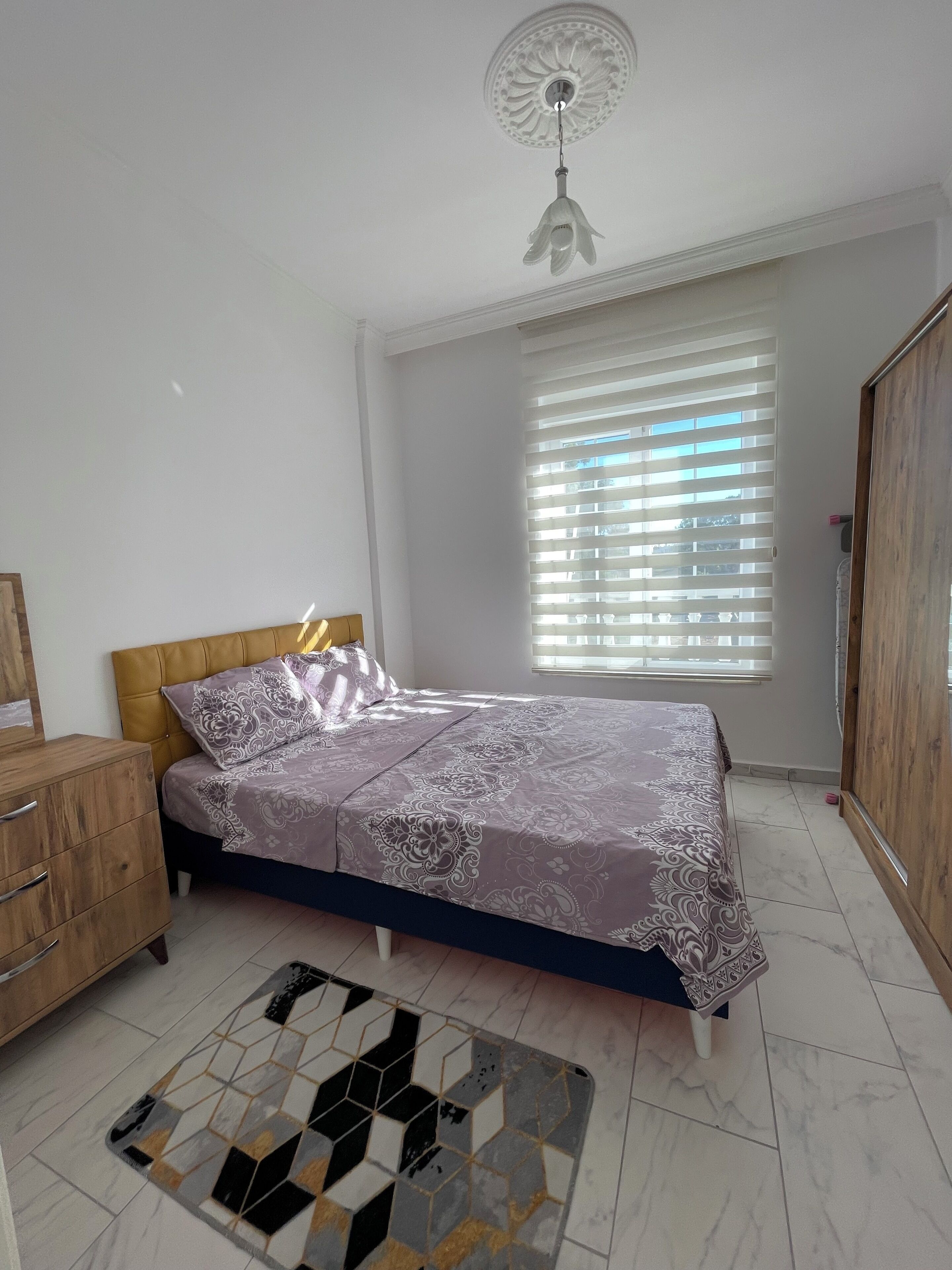 Ein Schlafzimmer mit einem Bett, einem Holzschrank, einem gemusterten Teppich, einem Deckenventilator und einem Fenster mit Jalousien.
