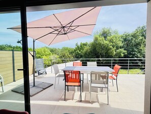 Een terras buiten met een tafel, stoelen, een grote parasol en uitzicht op bomen.