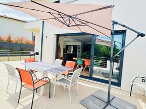 Un espace de restauration en plein air avec une table et des chaises, un grand parasol et une vue sur l’intérieur à travers une porte vitrée.