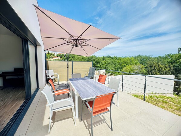 Une terrasse extérieure avec une table et des chaises, un grand parasol et une vue sur la verdure.