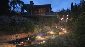 Une maison à plusieurs niveaux, dotée d’un mur de soutènement en pierre, d’une terrasse en bois et d’un éclairage extérieur.