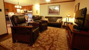 A living room with a sofa, a chair, a coffee table, and a television on a stand. There is a rug on the floor and a picture on the wall.