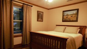 A bedroom with a wooden bed, a window with curtains, a framed picture on the wall, and a carpeted floor.
