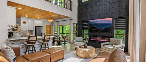 A spacious living area with a high ceiling, a kitchen in the background, and a large TV on the wall.