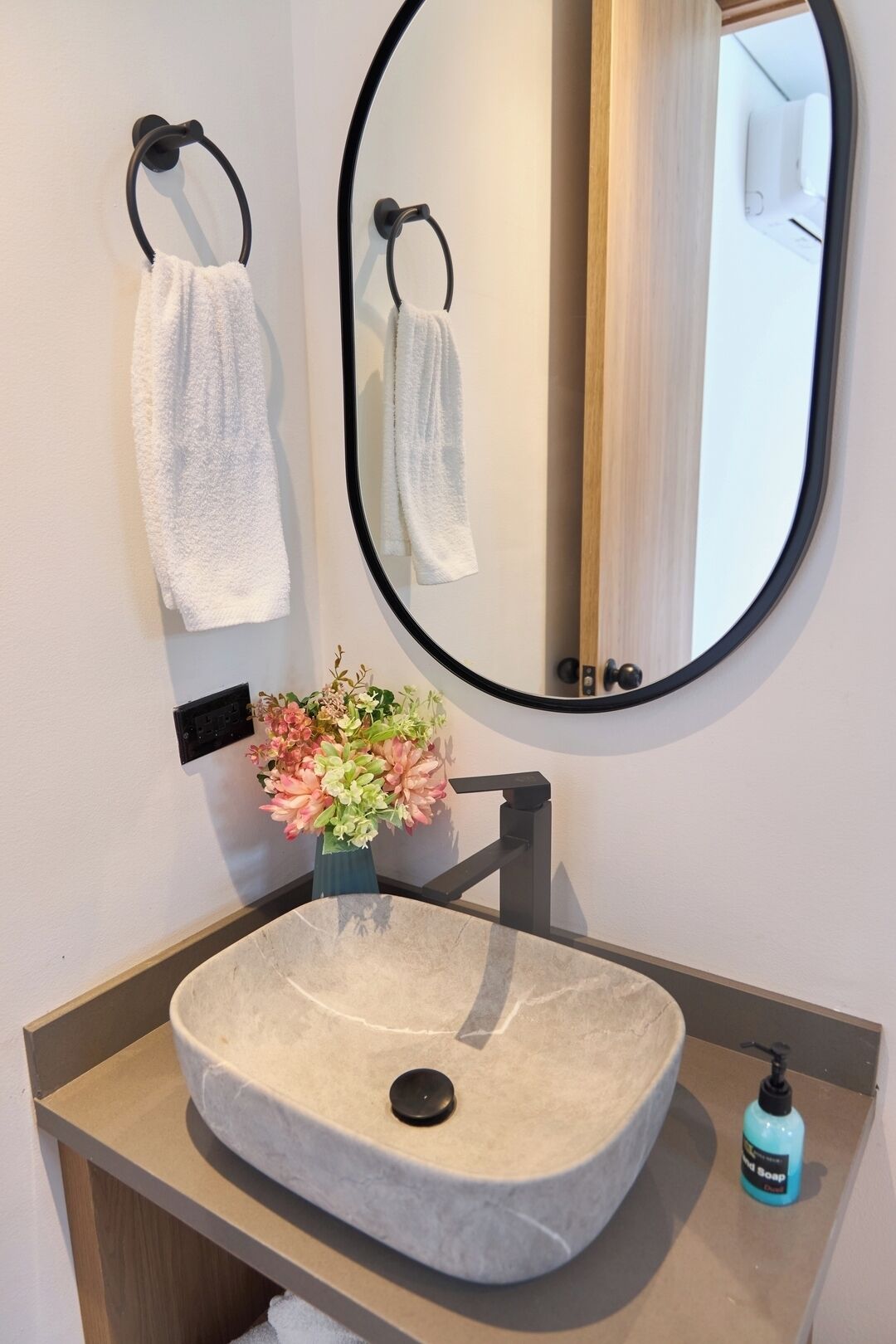 Une salle de bain avec un miroir rond, deux serviettes suspendues à des porte-serviettes, un vase avec des fleurs, un lavabo en marbre et un distributeur de savon.