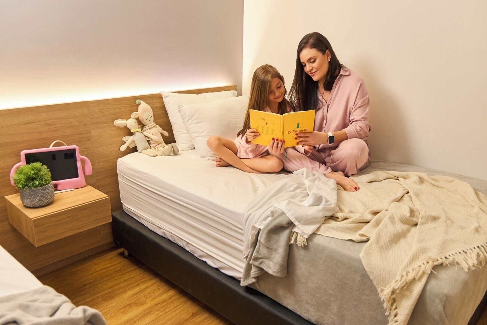 A bedroom with a bed, bedside table, and a nightstand with a pink tablet and a small plant.