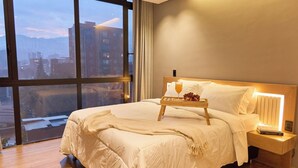 A hotel room with a large window, a neatly made bed, a bedside table with a lamp, and a tray with fruit and a glass of orange juice. - Stadio Haven Rosa Negra Cozy Suite (Medellín)