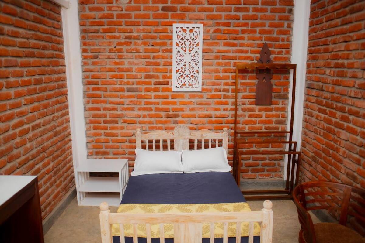 A room with a brick wall, a wooden bed with white and blue bedding, a small white bedside table, and a wooden chair.