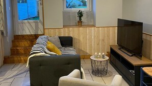 Ein Wohnzimmer mit einer Couch, einem Sessel, einem Fernseher und einem kleinen Tisch. Es gibt Fenster und einen Blick nach draußen.