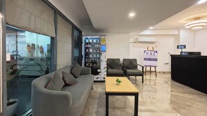 Een moderne lobby met een grijze bank, fauteuils, een bijzettafeltje, een automaat, een receptiebalie en een groot raam.