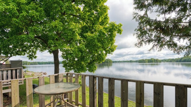Eine Holzterrasse mit einem Glastisch und Stühlen, mit Blick auf einen ruhigen See, wobei ein Baum im Vordergrund steht.