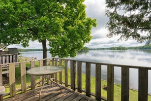 Une terrasse en bois avec une table et des chaises en verre, donnant sur un lac paisible, avec un arbre au premier plan.