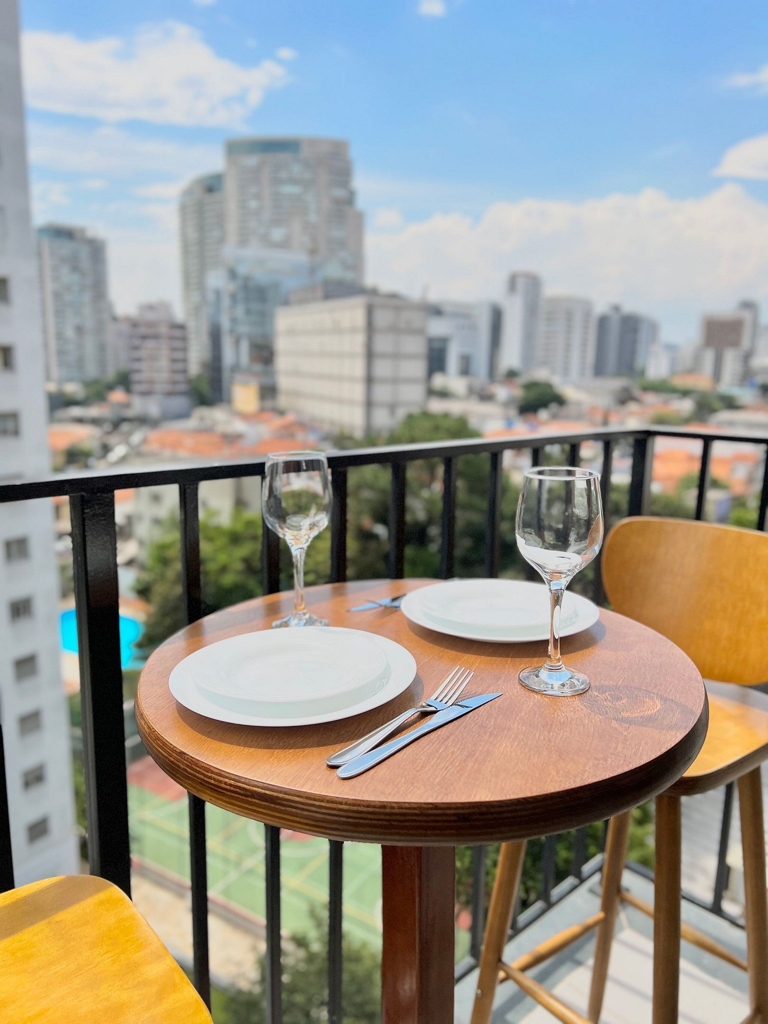 Mesa de madeira redonda com duas travessas brancas, um garfo e uma taça de vinho, dispostos numa varanda com vista para a cidade.