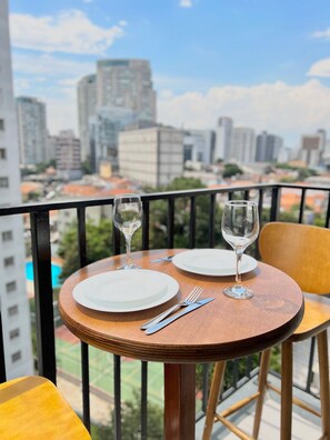 A round wooden table with two white plates, a fork, and a wine glass set on a balcony overlooking a cityscape. - Innvista - VibePinheiros (São Paulo)