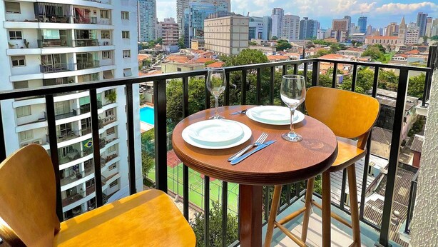 Un balcon avec une table dressée pour deux personnes, offrant une vue sur un paysage urbain avec de hauts immeubles et un ciel dégagé.