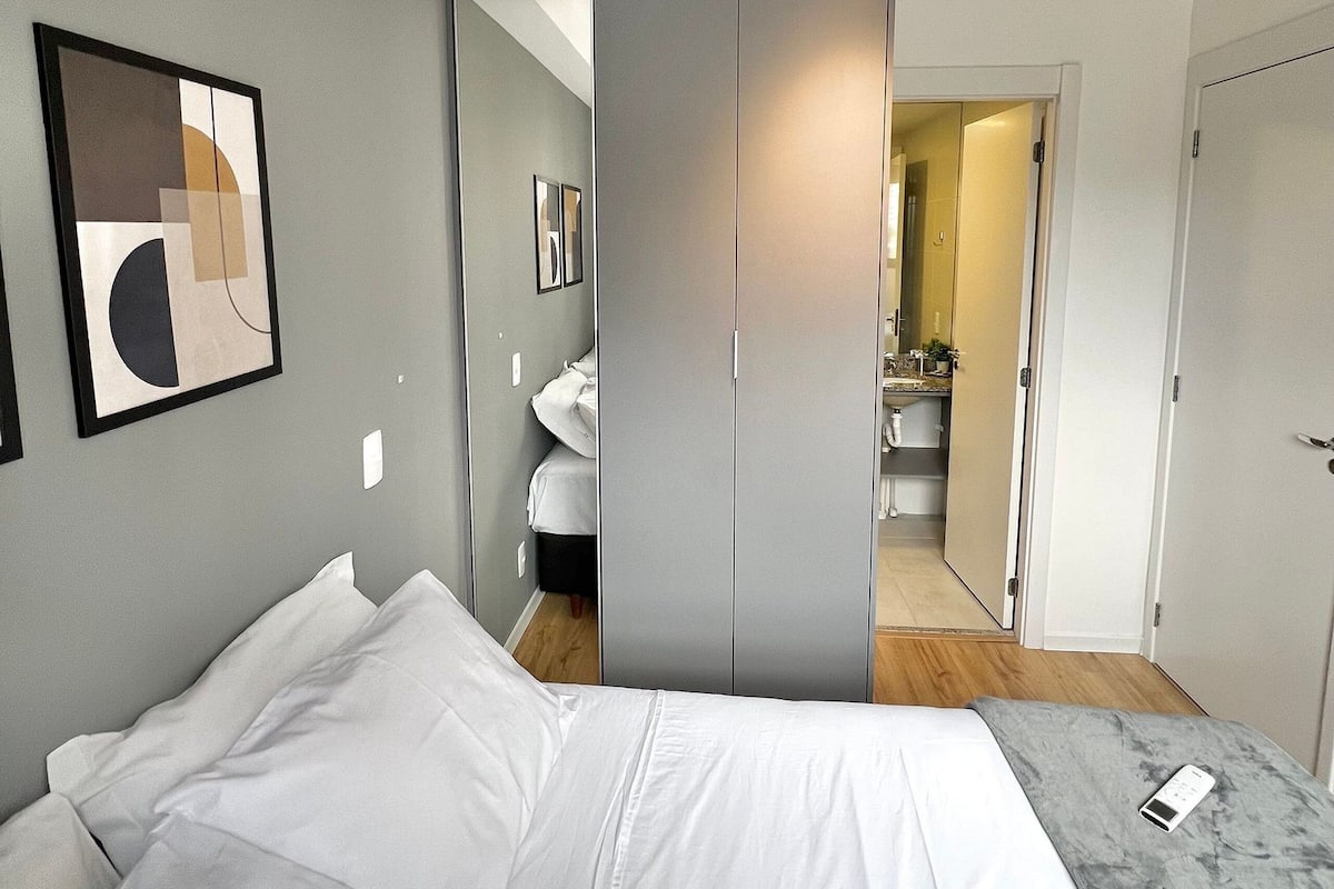A modern bedroom with a large bed, a grey wardrobe, a bathroom visible through a door, and a framed abstract artwork on the wall.