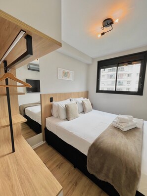 A modern hotel room with a large bed, a wooden headboard, a bedside table, a hanging light fixture, a window with a view of buildings, and a wall-mounted air conditioning unit. - Innvista - VibePinheiros (São Paulo)