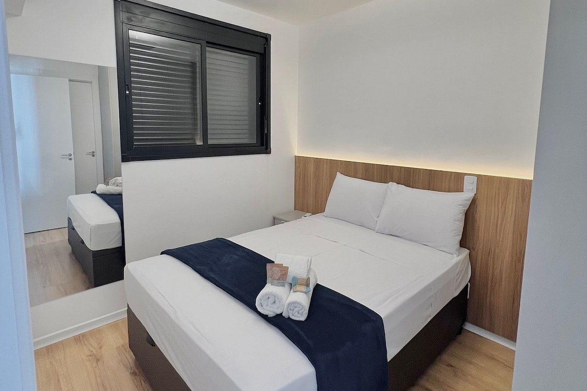 A modern hotel room with a single bed, a bedside table, a mirror, and a window with blinds.