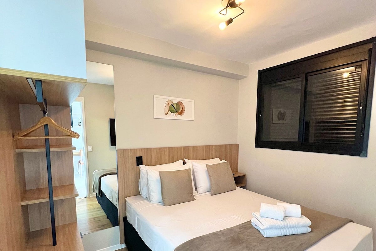 A modern hotel room with a large bed, a wooden wardrobe, a bedside table, and a window with blinds.