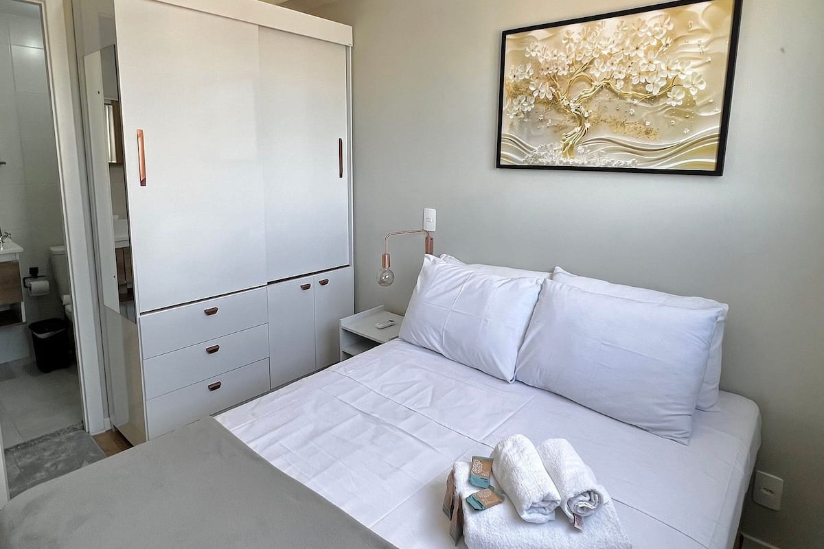 A hotel room with a bed, a wardrobe, a nightstand, a wall-mounted lamp, and a framed artwork.