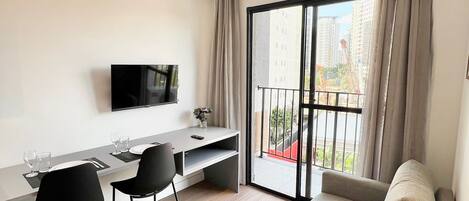 Un salon moderne comprenant un canapé, une table à manger avec des chaises, un téléviseur à écran plat fixé au mur et un balcon offrant une vue sur la ville.