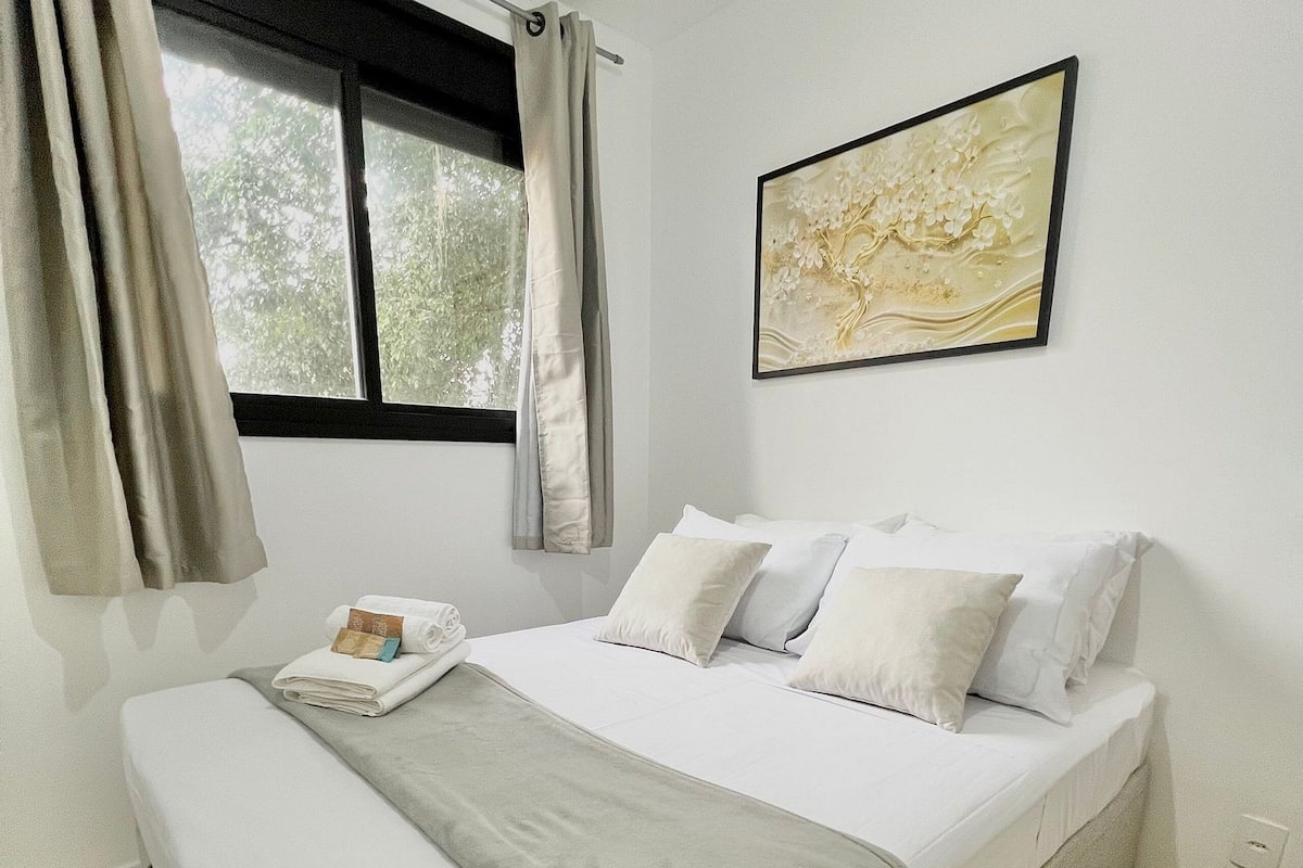 A neatly made bed with white bedding and a grey throw, a window with curtains, and a framed abstract painting on the wall.