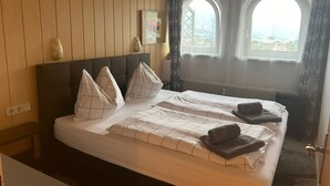 Una cama doble con una colcha a cuadros, dos almohadas y una toalla enrollada. La habitación tiene paneles de madera en las paredes y una ventana con cortinas.