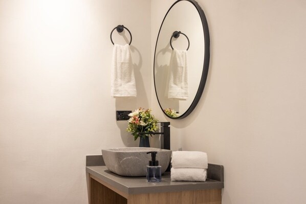 A bathroom with a round mirror, two towel rings, a sink with a vase of flowers, and a bottle of hand soap.