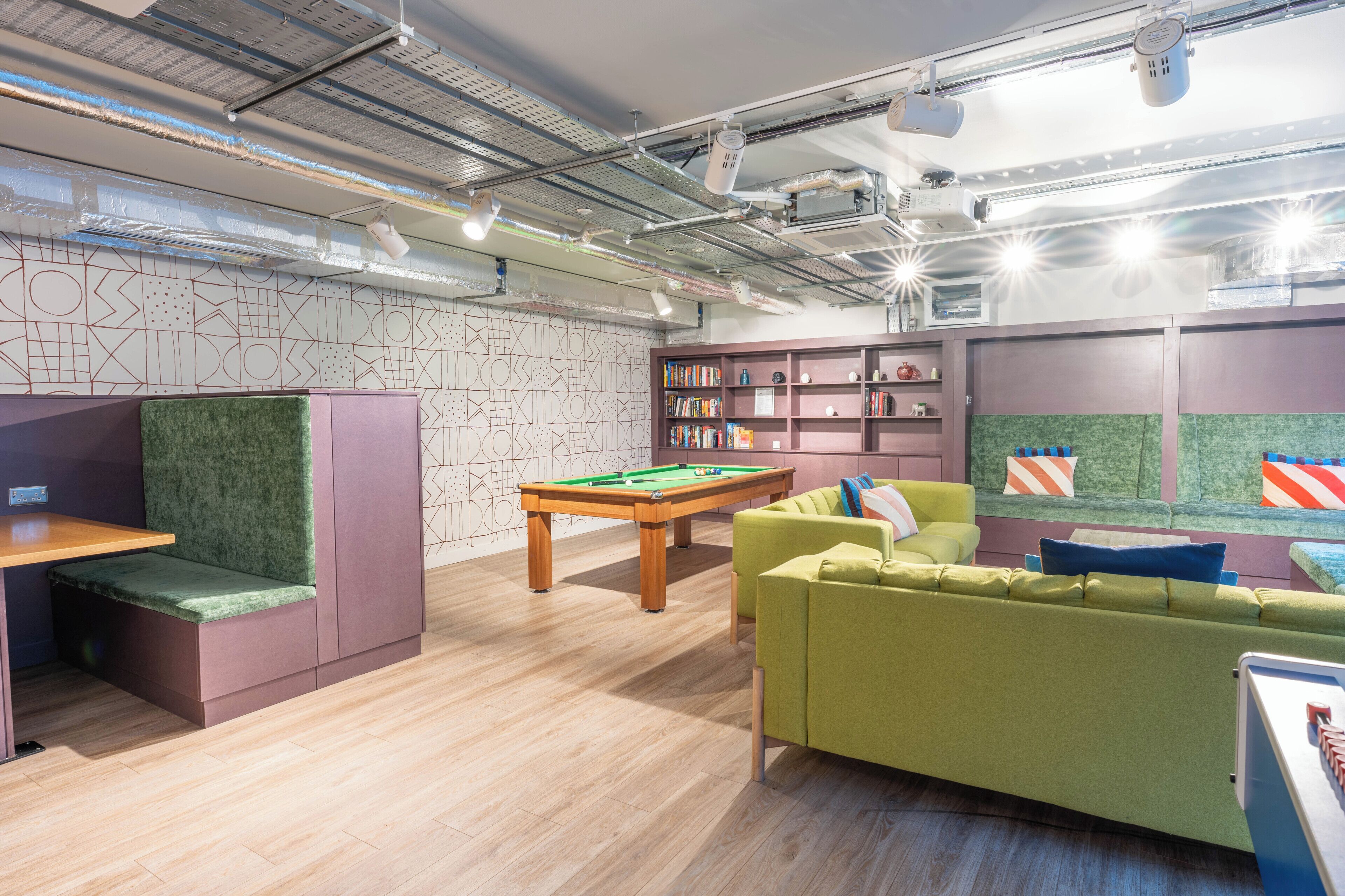 A modern lounge with a pool table, green cushioned seating, and a bookshelf.